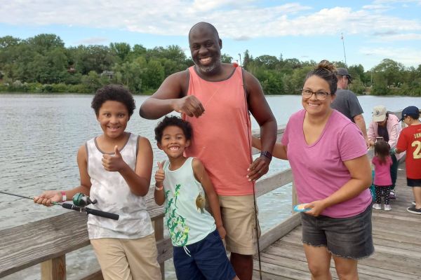 Two youths and two adults proudly hold up fishing poles as they stand on a wooden dock overlooking the lake on a partly sunny day. A forest surrounds the lakes’ borders in the background, with deep green trees lining the horizon. Everyone smiles for the picture, and the two youths are giving a thumbs-up sign.