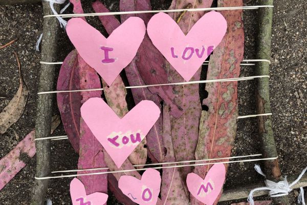 Four sticks are bound together by twine, creating a frame around pink-colored leaves and pink paper hearts that say “I LOVE YOU MOM” in a child’s handwriting.