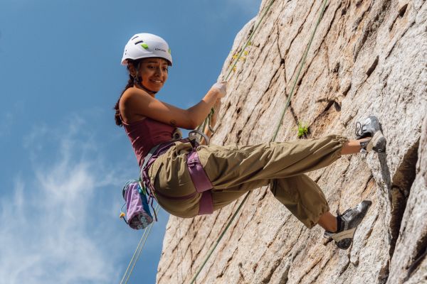 A girl, equipped with climbing gear, glances down and beams with pride as she scales a rocky slope that is completely vertical. Her body, which boasts a straight and confident posture, is supported by a harness that leads to the top of the slope. The sky above is a muted blue color, with wisps of clouds trailing by.