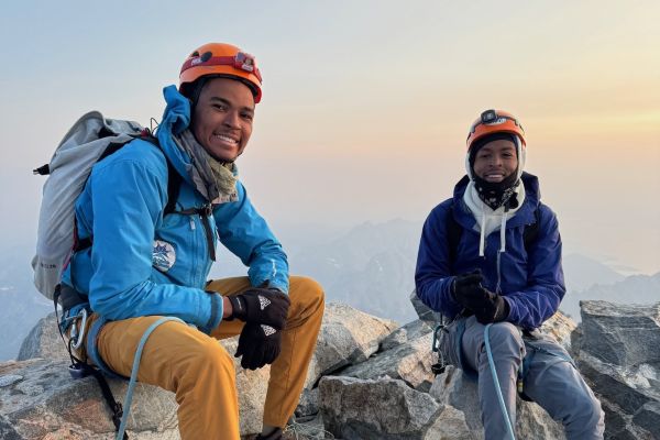 Two participants in climbing gear sit on top of stacks of large rocks to celebrate a successful climb in Grand Teton, Wyoming.