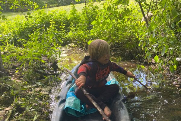 A young boy with blonde hair sits in a small plastic sled in a shallow stream surrounded by lush foliage. Holding a stick and wearing a red shirt and black pants he is enjoying an outdoor adventure.