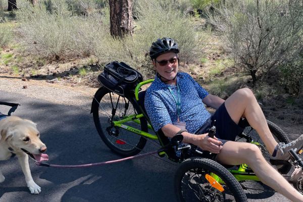 An Adventure Group participant rides a recumbent trike with his service dog on a sunny day. Behind their path are some trees and scrub, separating them from the main road.