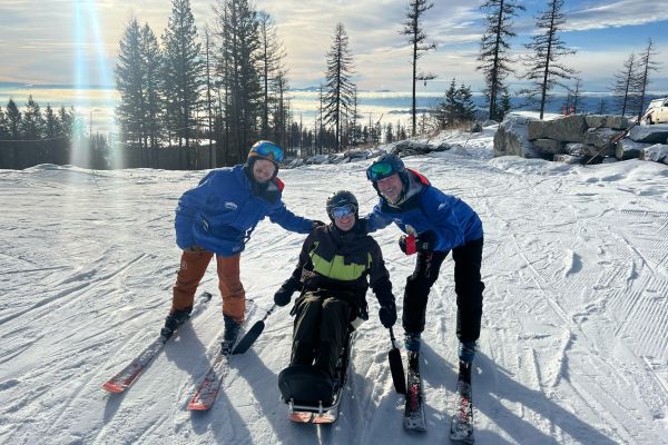 A participant is in a sit ski for a picture, with a DREAM volunteer and a seasonal staff employee posing on either side of her. The scenery behind them is an open ski slope covered with snow that shines bright from the sunlight.