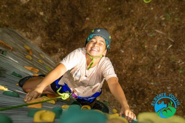 An overhead photo of an Elevate Youth member outdoor rock climbing at Hammond Pond looking toward the top of the route and smiling.