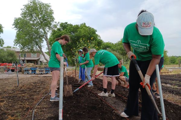 Seed Crew students diligently plow a row of soil in preparation for seed planting at Conundrum Farms. Lined up behind them are other already plowed rows. In the background are tall trees and a farm building; the sky above them is cloudy, which offers plenty of shade.