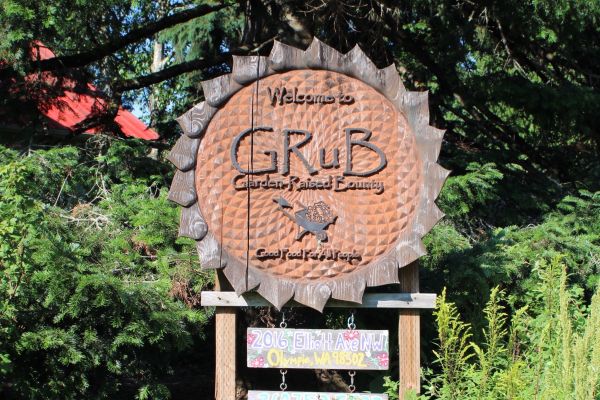 GRuB’s famous sign stands tall above thick, bright foliage. Its unique design reflects the shape of a sunflower, with GRuB’s logo placed proudly in the center. Behind the sign are rustling trees; a glimpse of a building’s orange rooftop can be seen between the leaves.