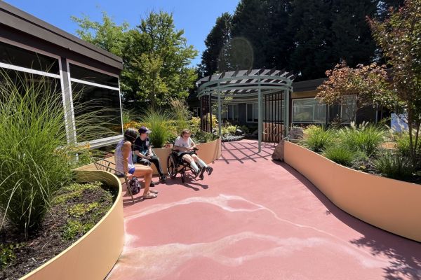 Three people—one of them in a wheelchair—are seated in a garden area that resembles an eddy, where fresh air is gathered; rows of planters border the walkway leading up to the seating area. The trio enjoys the spring bloom and the clear, sunny sky.