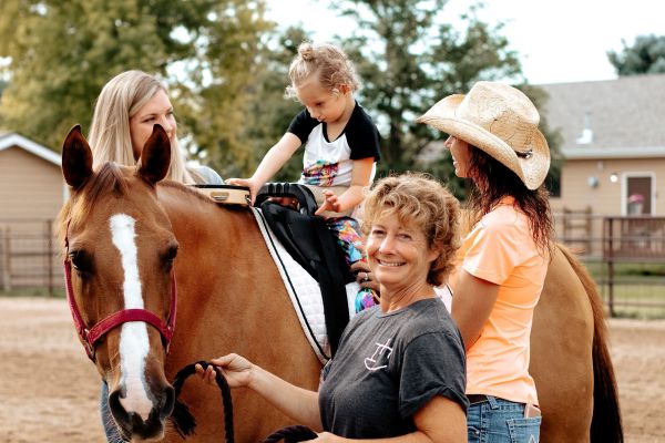 A young child is sitting on top of a horse with the support of three women. One woman is holding the reins, smiling warmly at the camera. The other two women stand beside the horse, helping the child in the saddle. The horse is brown with a white stripe down its muzzle. 