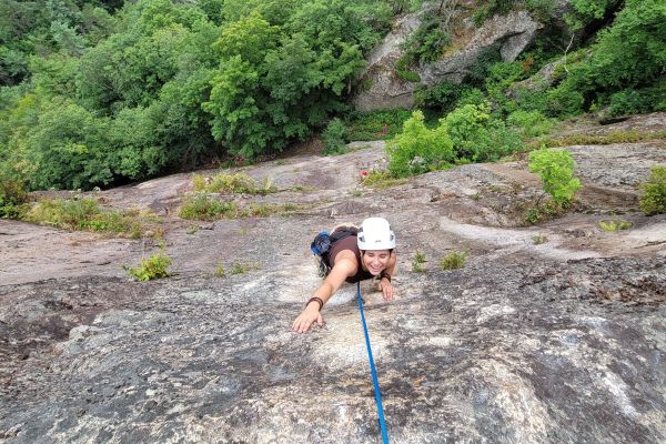 A young adult scales a steep rock face. The student is tethered by a rope and smiles at the camera with a sprawling forest below the cliff they are climbing.