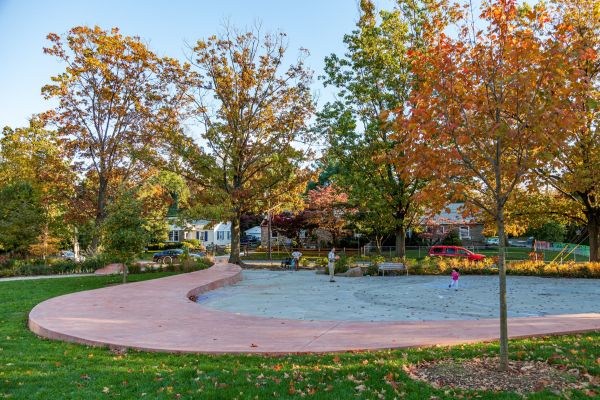 A salmon colored wall meanders through a park adjacent to lush green grass and open space. Several trees have leaves that are changing from green to orange.