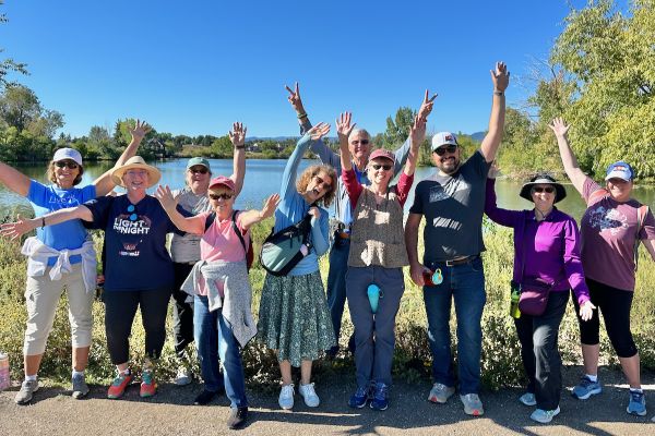 A group of participants pose with their arms up for a sunny picture in Belmar Park. Behind them is a vast lake surrounded by lush greenery.