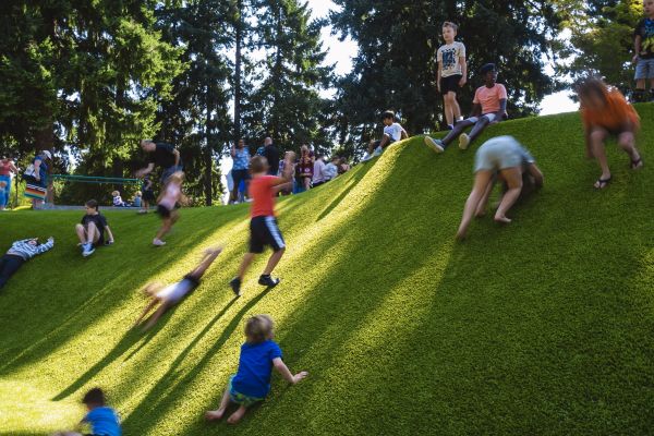 A crowded hill, full of playing children of all ages, blurred as the image captures them in motion. In the background, tall trees provide some shade from the sun.