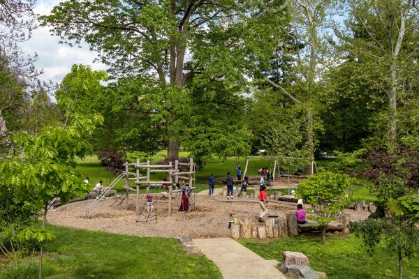 Multiple children are playing on an expansive wooden play structure surrounded by green grass and foliage.