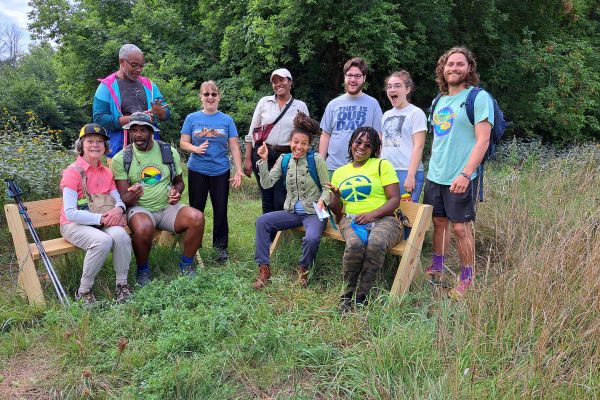 A group of participants gather closely to pose for a photo. Some people sit on wooden benches while others stand behind them. The scenery around them is full of green, as there are verdant trees behind them and tall wild grass surrounding them. The participants convey their admiration of the land through wide smiles.