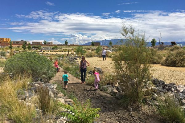 Children explore a muddy path made by a low-flow channel with their feet. One adult is watching the children play and another is walking through the adjacent field.