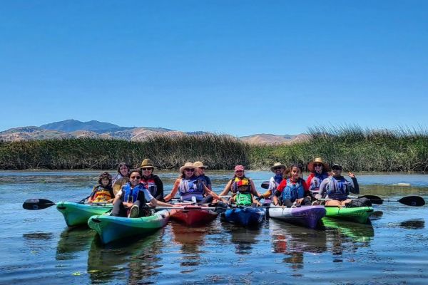 A group of people are kayaking down the CA Delta. The water itself is calm aside from the ripples caused by the kayaks. The blue sky above boasts no traces of clouds, promising a sunny day ahead for the participants.