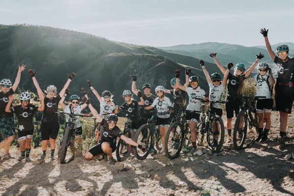 A large group of participants line up for a photo at the edge of a hill. They are all dressed in biking gear and have their arms thrown up in the air as a sign of triumph; some also have bikes by their sides. The background is a beautiful shot of a vast canyon, with rolling green hills that disappear far into the horizon. The clear sky overhead is lit up by a bright sun.