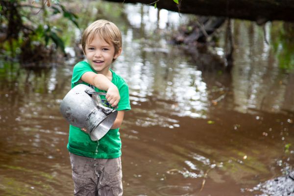 A young child stands smiling, ankle-deep in the shallow seasonal creek, holding a jug from which he pours. Behind him, a large log bridges the water, foliage draped across it.