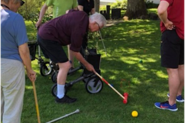 A group of people in summer clothes are standing with nearby ambulatory aids such as crutches, wheelchairs, and walkers. Two people are holding croquet mallets, with one leaning over and preparing to hit the yellow ball.