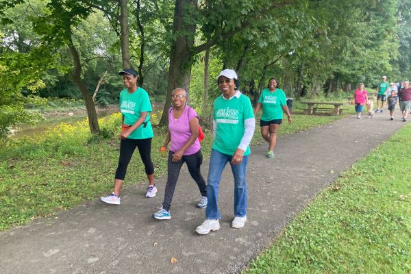 Multiple people are walking in small groups along a tree-lined path, many are wearing blue Walk with a Doc t-shirts, and all are smiling.