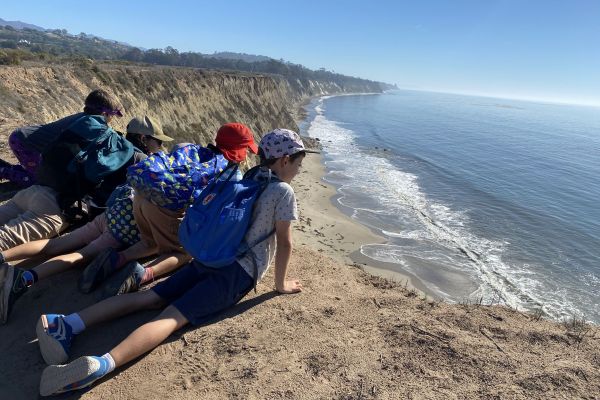 Four kids in hiking clothes lay by the edge of a cliff that overlooks the ocean. They eagerly admire the landscape spread before them, which includes a sunny, cloudless sky and a vividly blue ocean that disappears into the horizon.