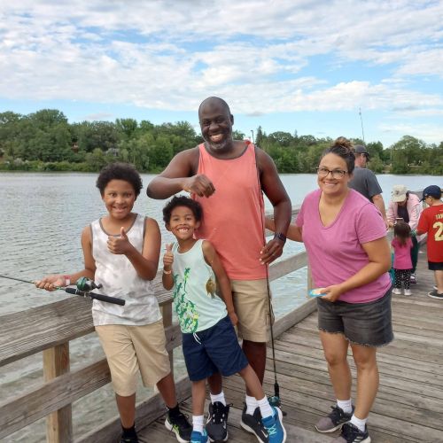 Two youths and two adults proudly hold up fishing poles as they stand on a wooden dock overlooking the lake on a partly sunny day. A forest surrounds the lakes’ borders in the background, with deep green trees lining the horizon. Everyone smiles for the picture, and the two youths are giving a thumbs-up sign.