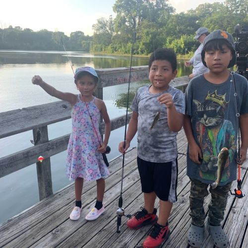 Standing on a wooden dock overlooking the lake, three children grasp their fishing poles, with two of them showing off the small fish they caught. In the background, a photographer peers at the children from behind their camera with a smile on their face.