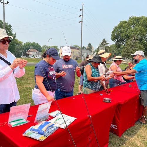 Under an overcast sky, a large group of adults wearing fishing hats stand around a long rectangular table covered with a  red tablecloth. On the table are fishing materials that the participants are using to learn how to bait hooks. The area they are in is lush with greenery, from the grass they stand on to the tall trees that frame the background.