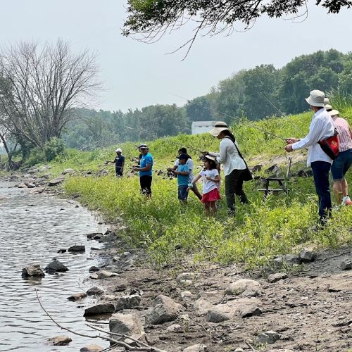 On an overcast day, a group of nine youths and adults stand in a line along the rocky, grassy bank of a lake. They cast their fishing poles and wait patiently for fish to bite. Some are fishing individually while some adults are paired with youth, instructing them on how to cast. Tall, verdant trees in the background can be seen from a distance.