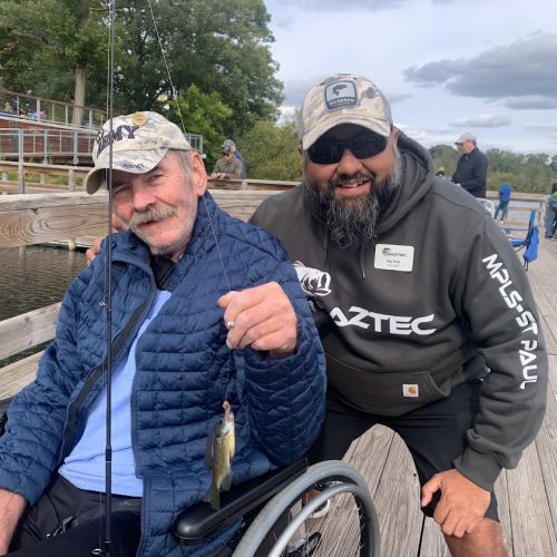 An older man in a wheelchair holds up a small fish and fishing pole, with a bearded Baztec staff member crouching behind the man and smiling in encouragement. They are both on a dock, with several people in the background gathered at the dock’s edge. The sky is overcast with clouds, but that doesn’t stop participants from enjoying the lake scenery. 