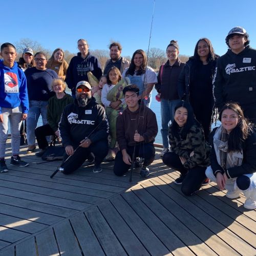 Under a clear blue and sunny sky, a group of youth and Baztec staff pose as they smile for a photo. They stand on a wood dock with the lake spanning out behind them; the orange and brown trees in the far distance show signs of the autumn season.