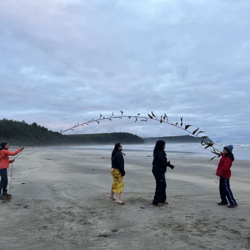 Four participants clothed in cold weather gear play jump rope with a long seaweed strand as they stand on a beach while on a camping trip on the coast of Washington. An overcast sky stretches over them, and in the background are a view of the misty ocean and patches of forested regions. 