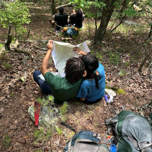 Four expedition participants take a break from their hike outside Birmingham, AL. They sit in pairs on a shaded, leaf-covered trail surrounded by vibrant green trees as they examine a trail map. They have taken off their backpacks and hiking gear to reorient to the trail.