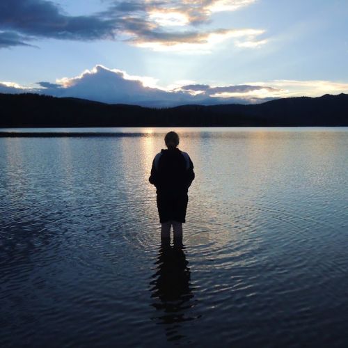 A youth participant wades in one of the lakes in the Rocky Mountains, his steps causing the water to ripple around him. He looks out towards the sunset horizon, which is reflected in the lake and colors the surface with pale orange hues.