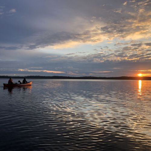 Two youth participants paddle in a canoe in the Boundary Waters of Minnesota and enjoy a vast view of an orange sun peeking over the cloudy horizon. The gradient sunset sky reflects onto the rippling water surrounding them.