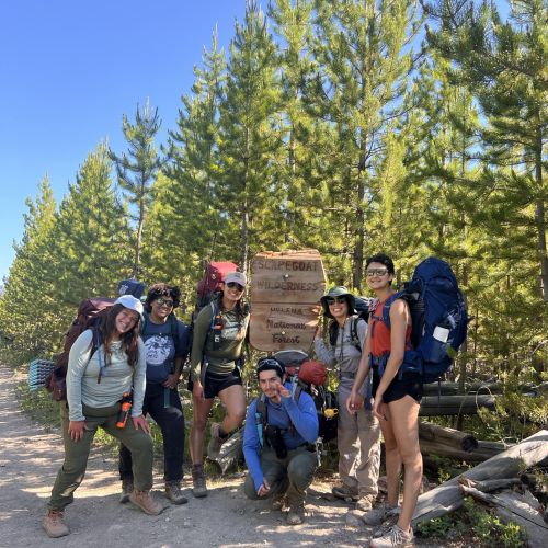 Volunteers carrying hiking gear on their backs pose in front of a wooden sign with bright smiles on their faces as part of Wilderness 101 – an education focused trip bringing BIPOC Wilderness visitors to the Bob to create a safe learning space within the community. Behind them are rows of bright green trees that travel far along into the horizon, with a bright blue and clear sky looming over the entire landscape.