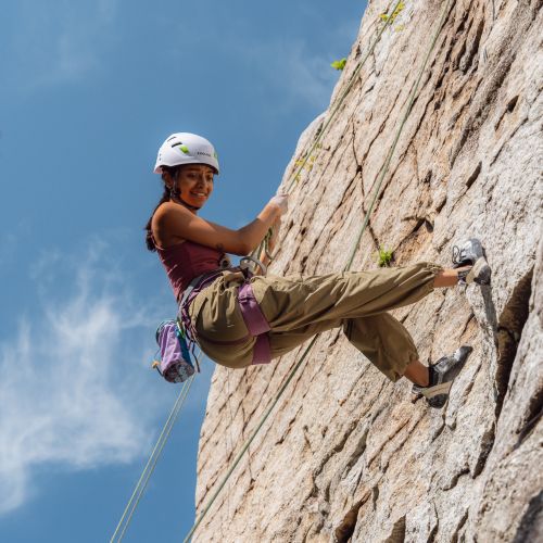 A girl, equipped with climbing gear, glances down and beams with pride as she scales a rocky slope that is completely vertical. Her body, which boasts a straight and confident posture, is supported by a harness that leads to the top of the slope. The sky above is a muted blue color, with wisps of clouds trailing by.