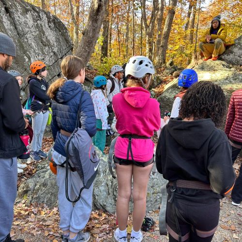 A group of climbers gather around a forest clearing to listen to a speaker, who is sitting on a large, sloped boulder. The woods around everyone are full of crisp orange and yellow leaves, and the ground itself is carpeted with fallen, brittle leaves—all of which spell the season of autumn.