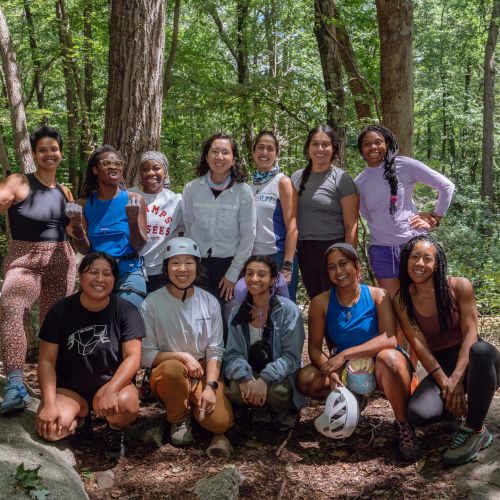 A large group of participants stand close under the dappled shade of a forest, which serves as a verdant background while they pose for a picture. Tall tree trunks jut from the ground behind the group, with leaves bursting from every angle that make up the dense foliage.