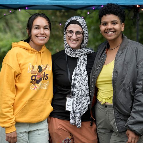 Three BGC participants pose together for a picture with bright smiles on their faces. Behind them is dense foliage and deep green trees, with beams of sunlight filtering through the tree canopy. Climbing equipment is set up behind them, though their bodies hide it from clear view of the camera.