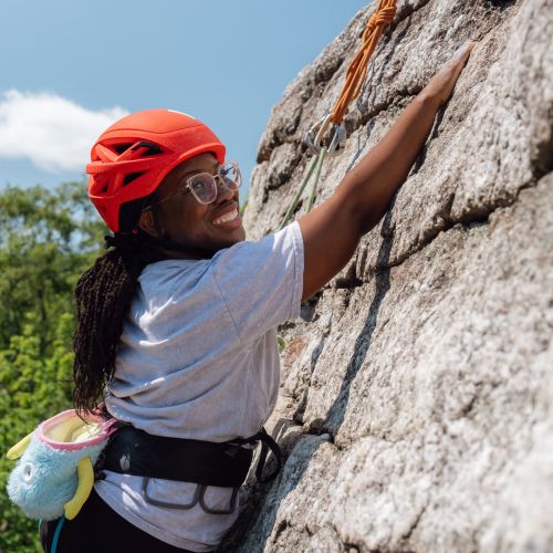  A climber in an orange helmet is in the middle of scaling a cliff and grips onto an edge in the rock surface. Though in the middle of climbing, she throws a bright smile at the camera. In the distance are the tops of bright green trees and a bright blue sky stretching overhead.