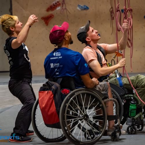 A climber who uses a wheelchair belays another climber during an adaptive climbing program in Murfreesboro, TN. Another person in a wheelchair is watching the belayer and climber on the climbing wall. A volunteer is standing behind the belayer with her arms raised in the air. Ropes hang from the climbing wall in front of the belayer in his wheelchair. 