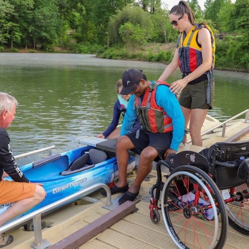 A kayaker transfers from his wheelchair into the kayak using the accessible dock and transfer bench while two volunteers help stabilize the kayak. A third volunteer stands behind the adaptive kayaker with her arms on the adaptive kayaker’s shoulders. The adaptive kayaker and volunteer are on a wood dock next to the riverbank. Several yellow and orange kayaks are stacked on top of each in the water at the end of the dock. The river recedes into the distance lined with leafy green trees. 