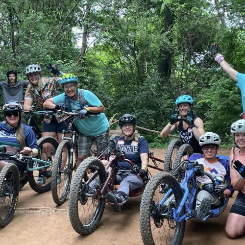 A group of adaptive mountain bike participants stop for a photo with a team of volunteers on bikes behind them, during the AMBC Fall Fest in Knoxville, TN. The adaptive riders are seated on their bikes smiling. Several volunteers are leaning against their bikes on the dirt bike path in front of leafy green trees. 