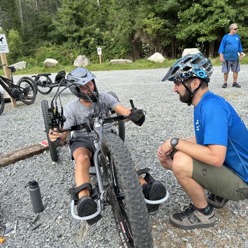 Adaptive Mountain Bike director, Matt Kirby, kneels on the ground while helping to prepare a rider on his bike at an adaptive mountain bike ride in Asheville, NC. The two are surrounded by mountain bikes and other volunteers standing near the edge of the woods. 