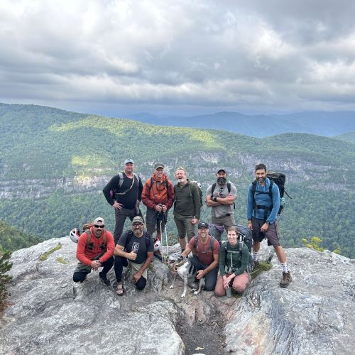 A group of veterans stop for a group photo at the summit of Table Rock in North Carolina during the 9/11 Tribute climb that takes place every year. The veterans are standing atop the granite dome of Table Rock smiling at the camera. In the background, the hazy rolling hills of the Great Smokies recede into the distance under a blue sky filled with fluffy white clouds. 