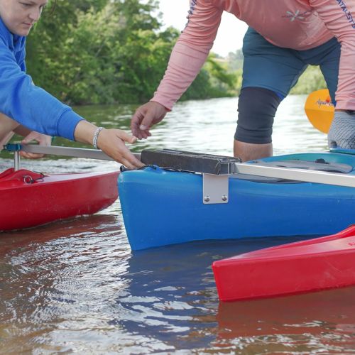 Volunteers help to attach the outriggers on a kayak. Two Adaptive Kayaking program volunteers stand in the water and stabilize the kayak. In the background, the river stretches into the distance. The riverbank is lined with leafy green trees. 