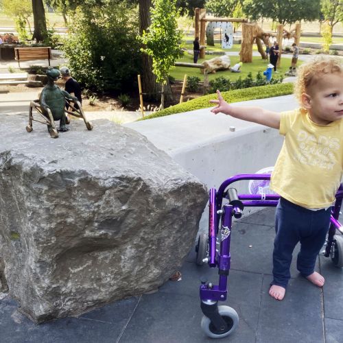 A toddler with a walker is pointing to the bronze statue of a turtle, also using a walker, on a sunny day in the playground.