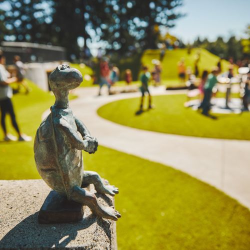 A bronze turtle statue sits on a stone in the foreground as families play in the busy park on a sunny day.