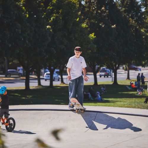 A small child with a helmet on a bicycle looks up to a skateboarding teen on a concrete bowl. The CHAMPS wheel play zone is in front of a shaded park full of families.